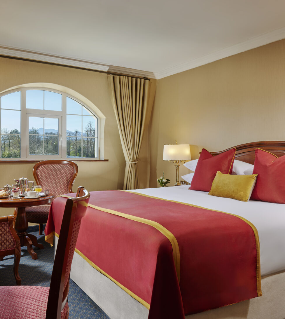 Bright hotel room with double bed, luxurious red and gold accents, seating area, garden view, and elegant lighting.