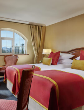 Elegant hotel room with plush red accents, scenic window view, cosy seating area, and warm lighting perfect for relaxing stays.