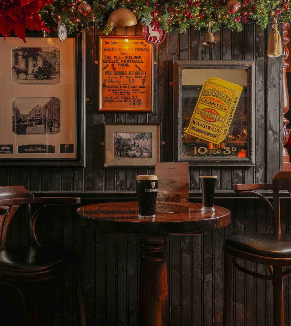 Cosy hotel bar with festive decor, vintage posters, wooden stools, and a table with two pints, inviting atmosphere.