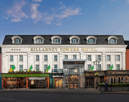 Elegant Killarney hotel facade with classic architecture, vibrant shops, and inviting entrance under a blue sky.
