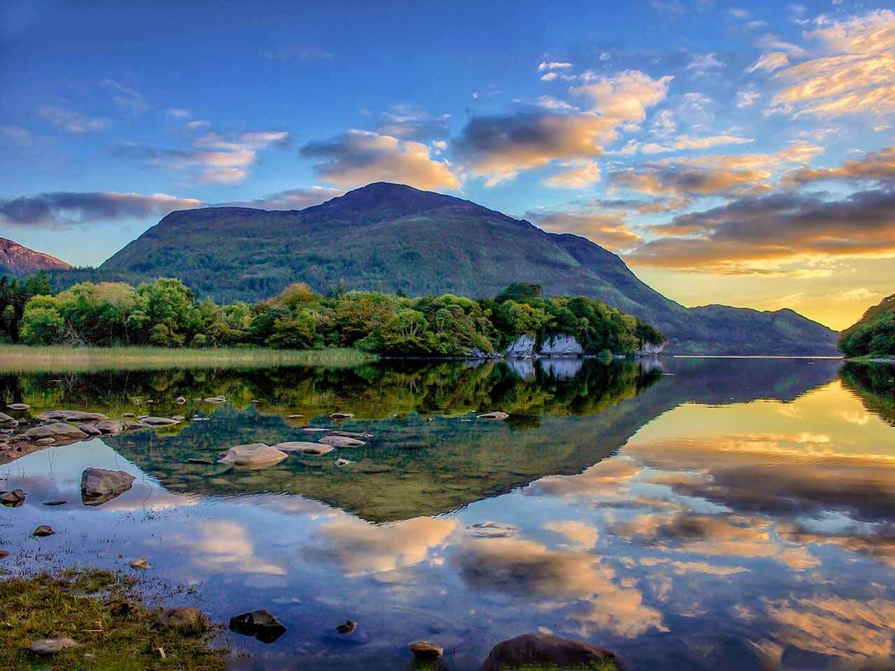 Serene lake with mountain reflection, lush greenery, and vibrant sunset skies, inviting relaxation and nature exploration.