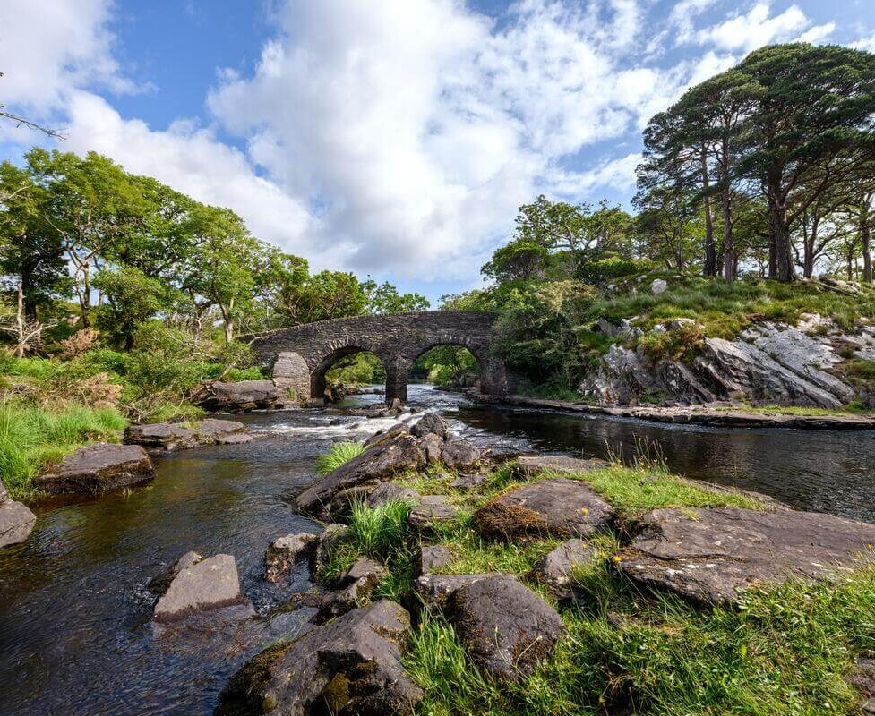 Picturesque stone bridge over tranquil river surrounded by lush greenery and blue sky at scenic hotel location.