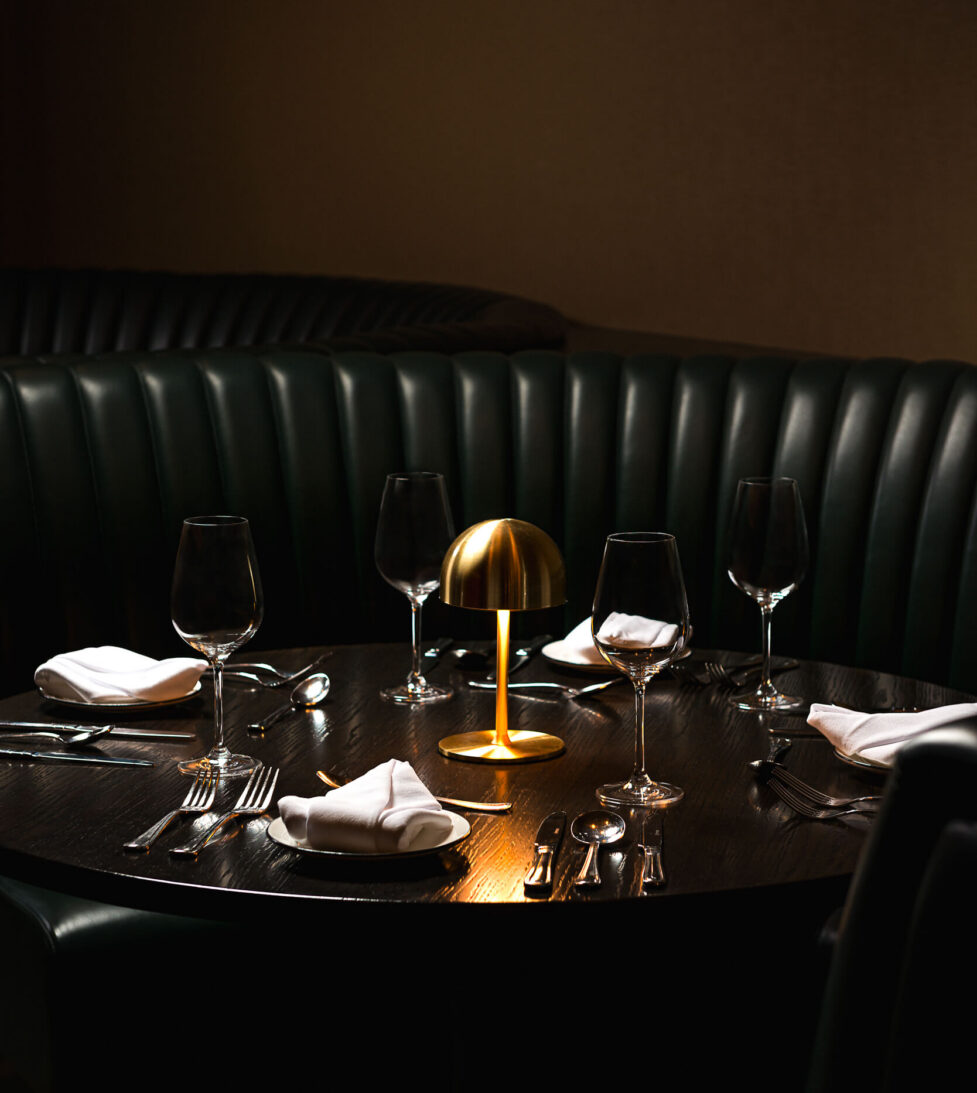 Elegant, dimly lit hotel restaurant with a cosy table set for four, featuring a gold lamp and plush green seating.
