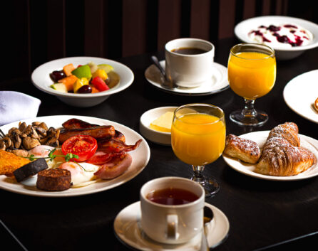 Full English breakfast with fresh pastries, fruit salad, and orange juice on a dark wooden table at cosy hotel dining.