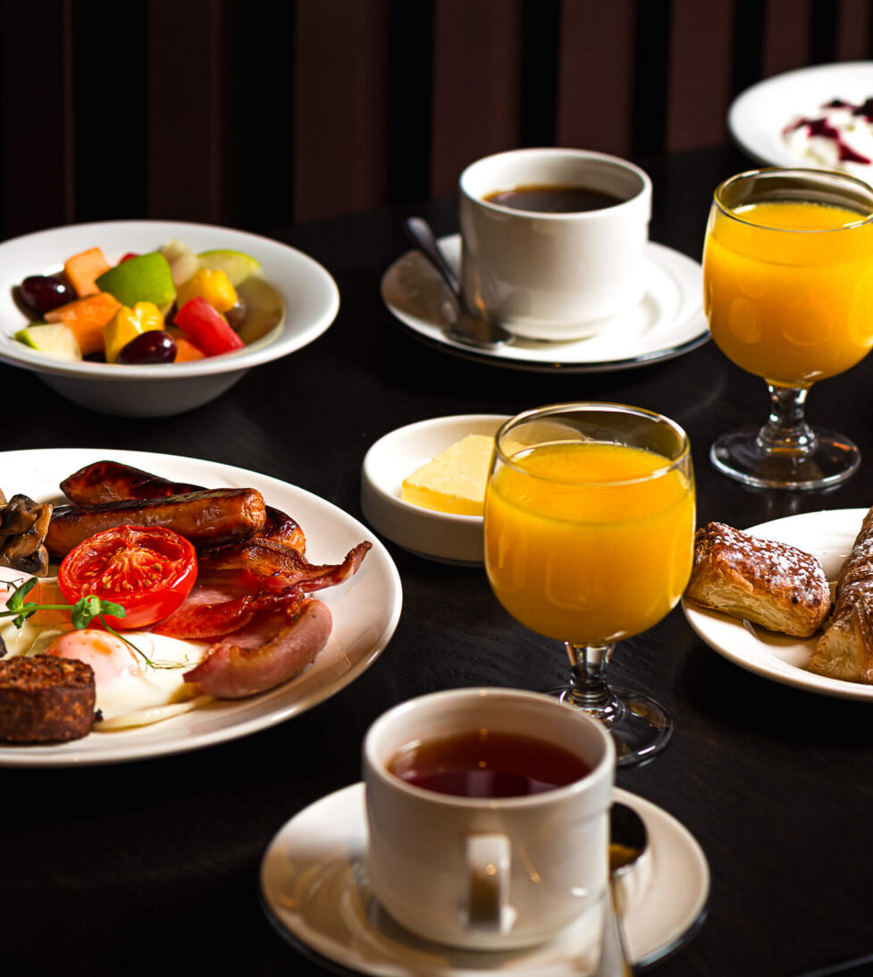 Full English breakfast with fresh pastries, fruit salad, and orange juice on a dark wooden table at cosy hotel dining.