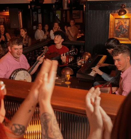 Lively hotel pub session with live band (banjo, guitar, keyboard), guests clapping, warm wooden interior and cosy table lamps.