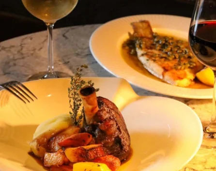 Slow‑roasted lamb shank with mashed potato, glazed root veg and herb garnish, served with white and red wine on marble table — fine dining hotel restaurant