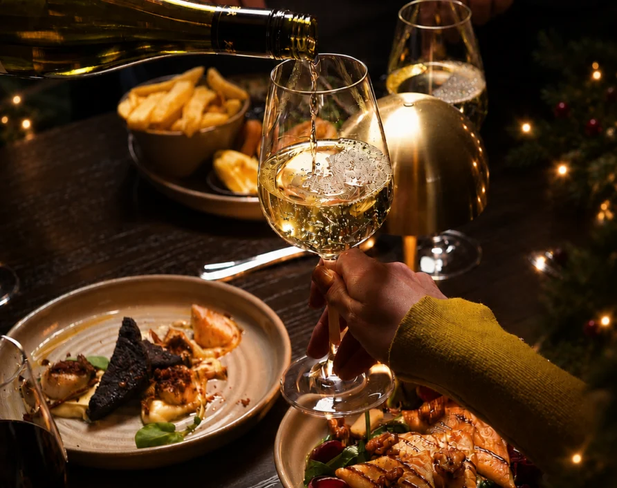 Festive hotel dinner table with white wine being poured, plated seafood and salad, fries, candlelight and cosy ambient lighting